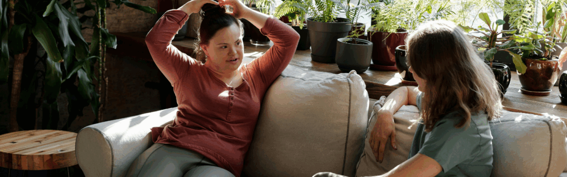 Kaksi naista, joista toisella on Downin syndrooma, keskustelevat. He istuvat sohvalla ja taustalla on kasveja.